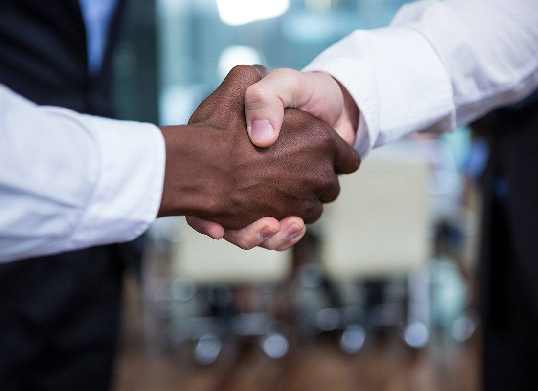 Employee Benefits Partners - Close Up of Two Professionals Shaking Hands in an Office with People Standing Around Them